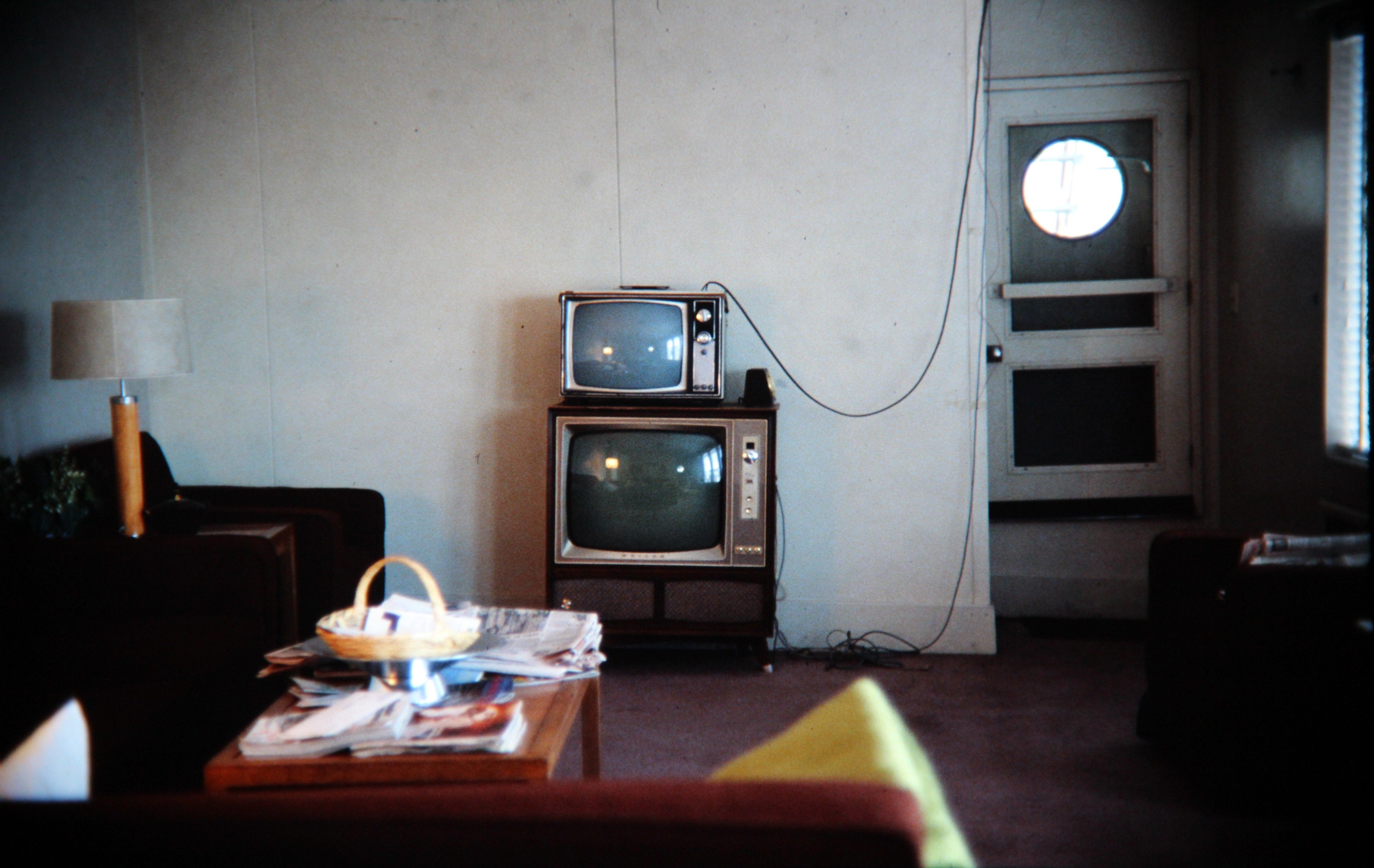 Picture of a sparcely-decorated living room. The walls are a plain white. There are two CRT televisions stacked on top of each other against the wall. There is a coffee table and chairs in the foreground and side out of focus.
