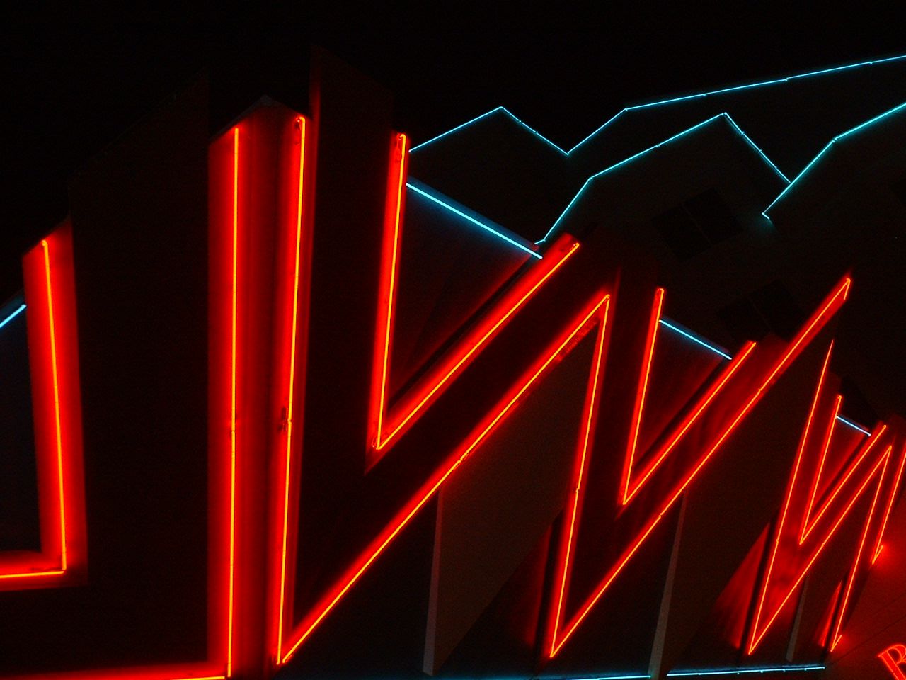 Close-up picture of red neon lights set at harsh angles. A set of blue neon lights resembling mountains is in the background.
