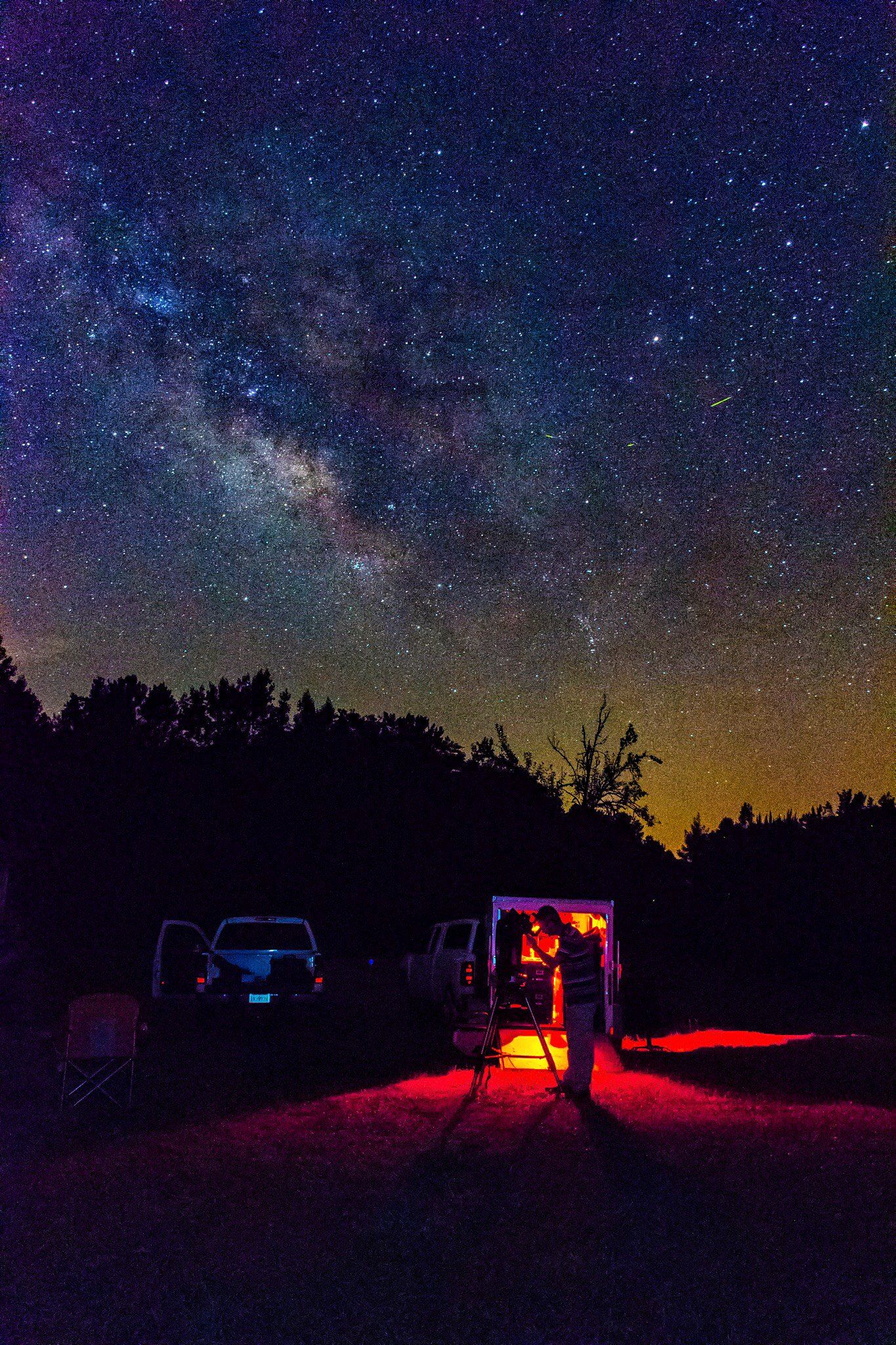 A man looking into a telescope backlit by red light coming from his van. The Milky Way and a shooting star are visible in the night sky.