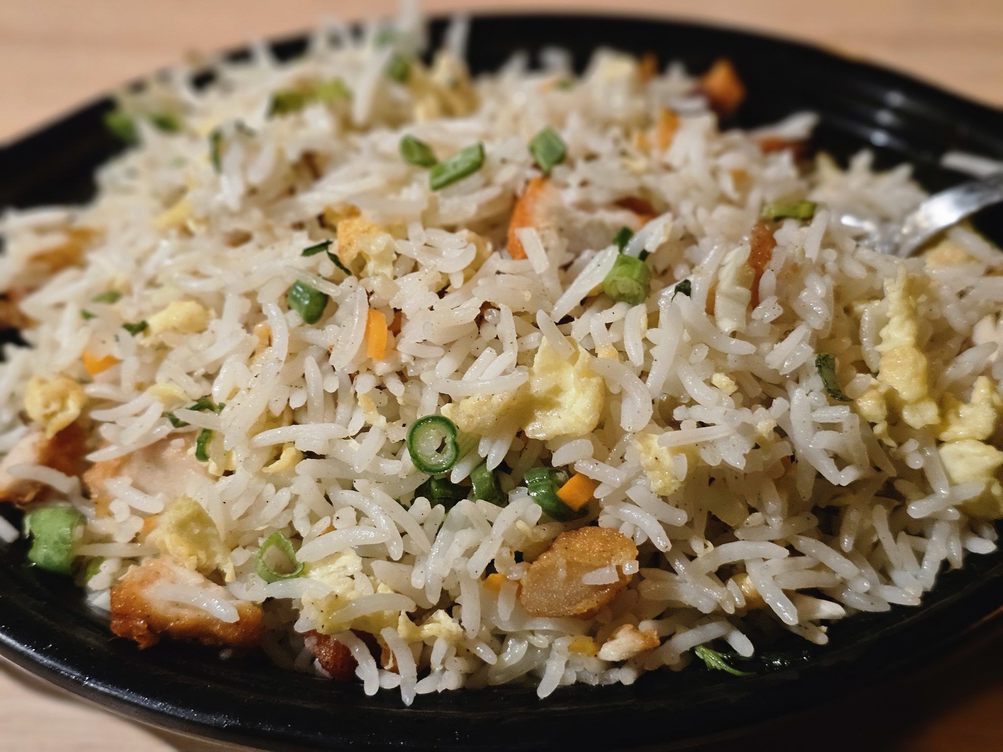 A close-up of a serving of chicken fried rice, featuring fluffy white rice mixed with diced carrots, scrambled eggs, and pieces of chicken, garnished with chopped green onions. The dish is presented in a black bowl, with a slightly blurred background emphasizing the texture and ingredients of the fried rice. It was captured in Kozhikode, Kerala.
